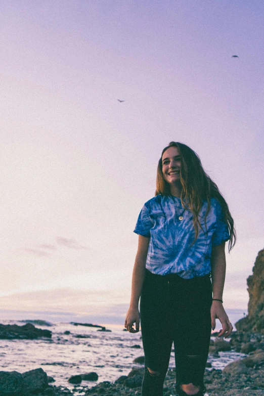 a woman standing on a rocky beach next to the ocean, unsplash, happening, wearing a tie-dye shirt, concert, smiling down from above, dusk sky
