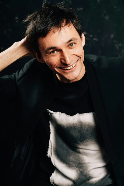 a man sitting on a couch with his hand on his head, an album cover, inspired by Anton Fadeev, antipodeans, while smiling for a photograph, standing with a black background, marc _ simonetti, headshot profile picture