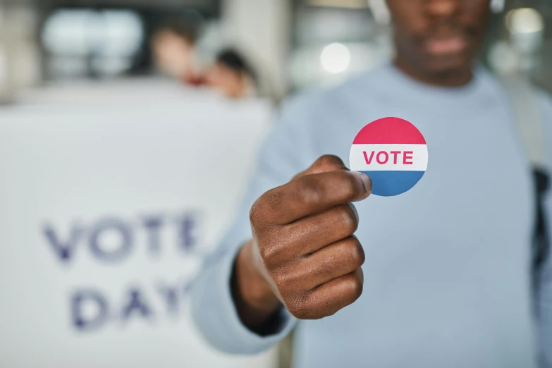 a man holding a sticker with the word vote on it, a photo, by Matija Jama, shutterstock, renaissance, good day, instagram story, no - text no - logo, a person standing in front of a