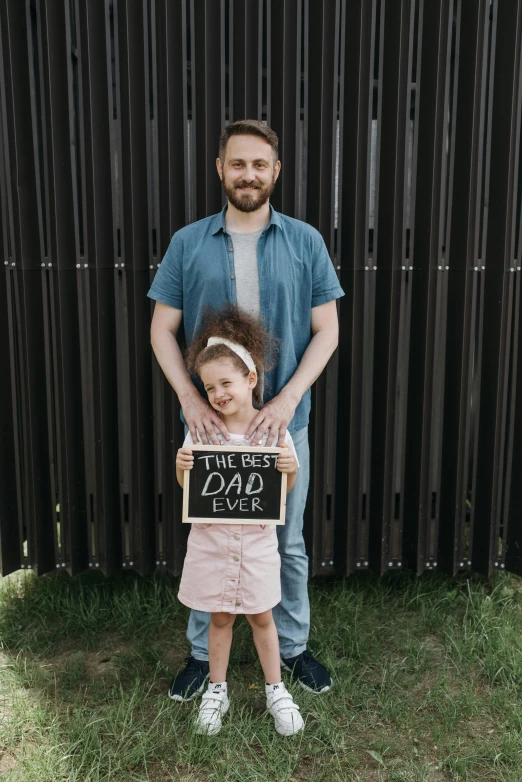 a man standing next to a little girl holding a sign, an album cover, pexels contest winner, with a beard and a black shirt, third trimester, chalkboard, instagram story