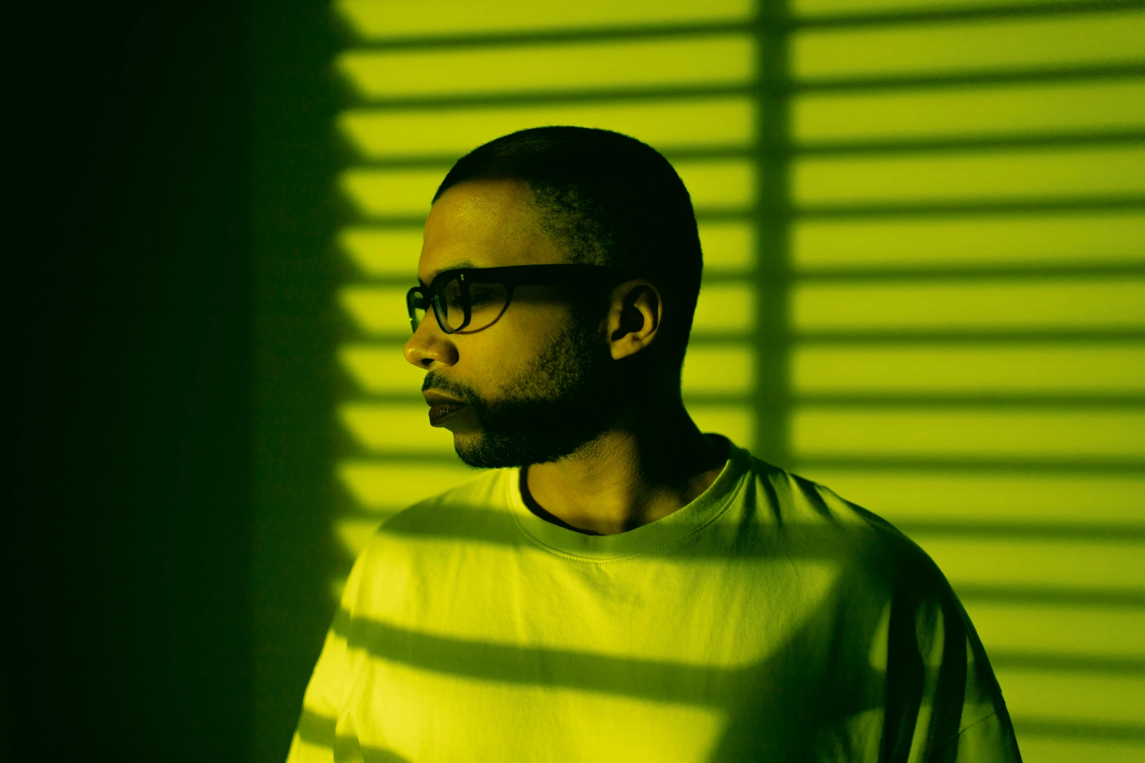 a man with glasses standing in front of a window, an album cover, inspired by Liam Wong, pexels, hurufiyya, yellow infrared, green lights, ((portrait)), malcolm hart