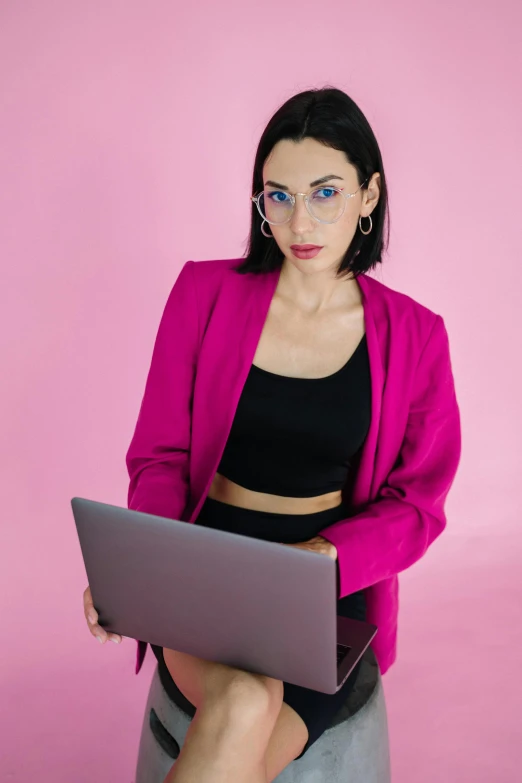 a woman sitting on a stool with a laptop, by Julia Pishtar, trending on pexels, wearing a pink tux, with square glasses, mia khalifa, androgynous male