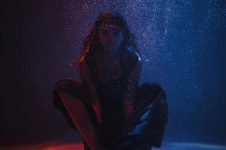 a woman sitting on the ground in the rain, an album cover, unsplash contest winner, holography, red and blue black light, water particles, madison beer as leeloo, dark. studio lighting
