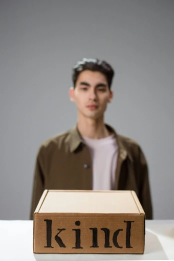 a man standing in front of a box with the word kind on it, an album cover, inspired by Fei Danxu, trending on unsplash, hyperrealism, non binary model, made of cardboard, headshot profile picture, carrying a tray