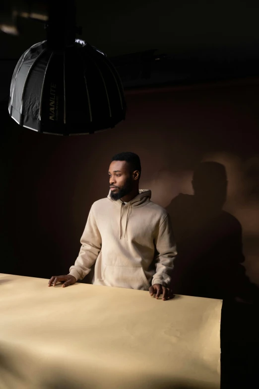 a man standing next to a table in a dark room, an album cover, inspired by Gordon Parks, photorealism, he is wearing a brown sweater, pencil drawing of mkbhd, dramatic soft shadow lighting, photo from a promo shoot