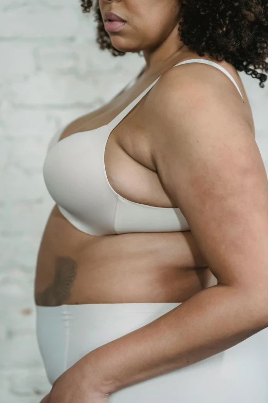 a woman in a white bra top standing in front of a brick wall, detailed product image, showing curves, anatomically incorrect, on a gray background