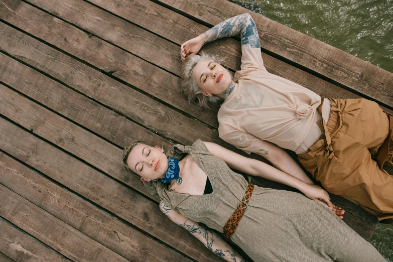 a couple of women laying on top of a wooden dock, a portrait, inspired by Elsa Bleda, trending on pexels, aestheticism, tattooed, pale hair, patterned clothing, nonbinary model