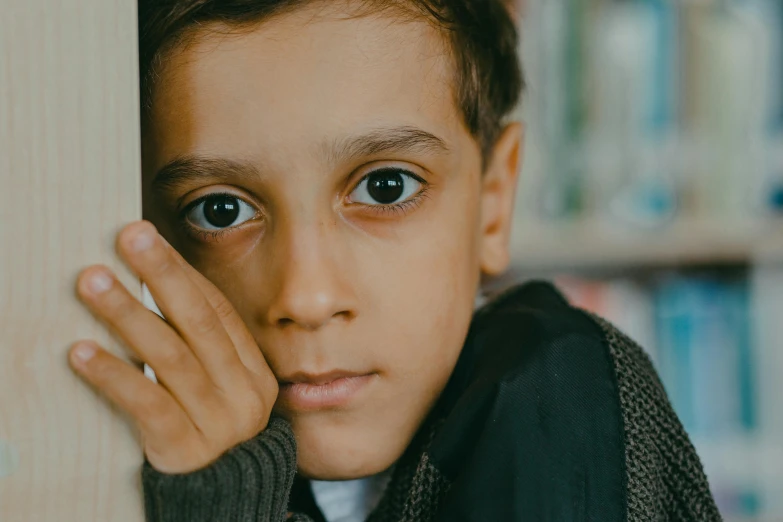 a young boy leaning against a wall in a library, pexels contest winner, hurufiyya, serious sad look in his eyes, brown hair and large eyes, 15081959 21121991 01012000 4k, lachlan bailey