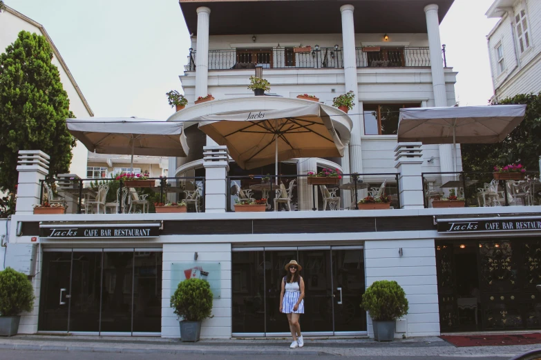 a woman standing in front of a white building, standing in a restaurant, meni chatzipanagiotou, balconies, family friendly