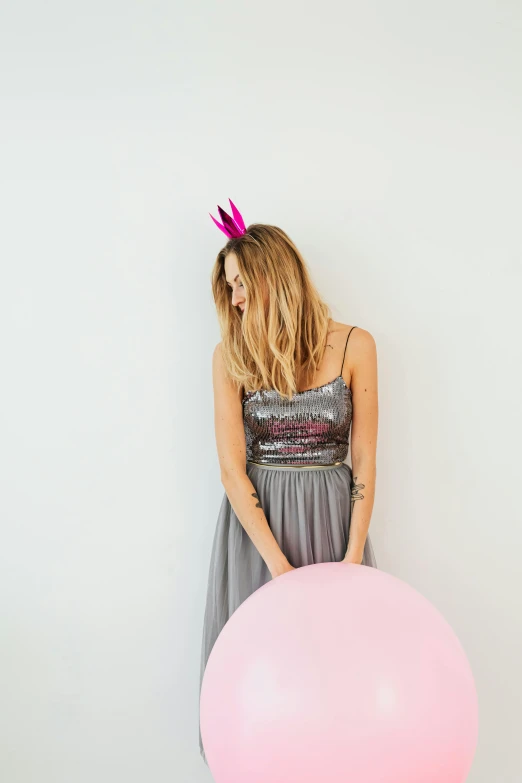 a woman holding a pink balloon in front of a white wall, wearing a light grey crown, party, dark. no text, grey