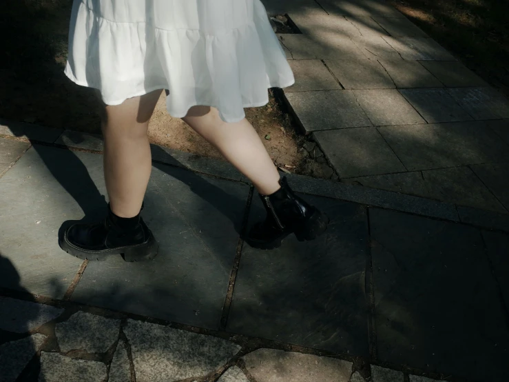 a woman in a white dress walking down a sidewalk, unsplash, realism, wearing black boots, extremely pale, in the shadows, walking at the garden