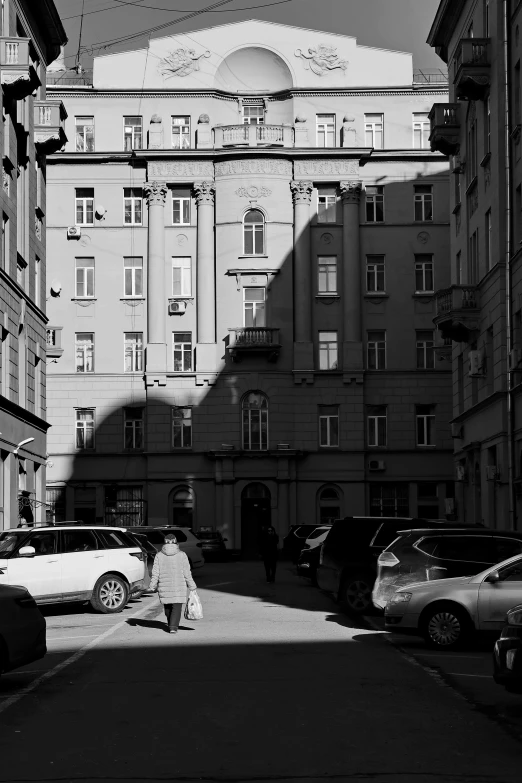 a black and white photo of a woman walking down a street, precisionism, gta in moscow, late afternoon sun, old buildings, parking in the street