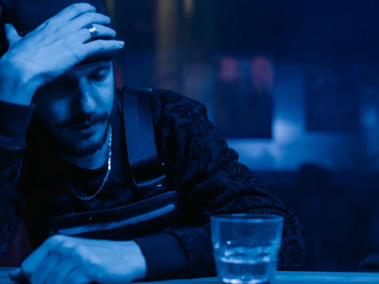 a man sitting at a table with a glass of water, inspired by Elsa Bleda, trending on pexels, renaissance, dark blue neon light, disappointed, shia labeouf, in a nightclub