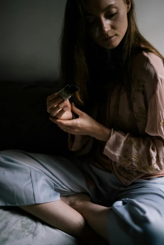 a woman sitting on a bed looking at her cell phone, inspired by Elsa Bleda, trending on pexels, holds a small knife in hand, nightgown, moody muted colors, carrying a bottle of perfume