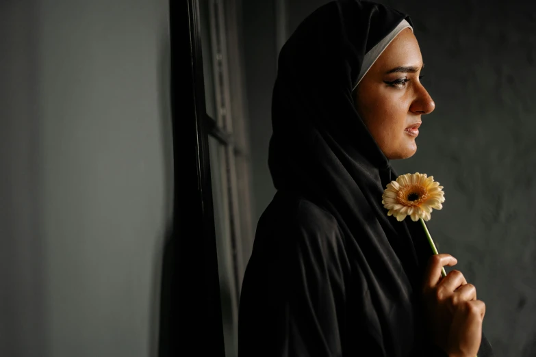 a woman in a hijab holding a flower, a portrait, pexels contest winner, looking out a window, lit from the side, black, over-shoulder shot