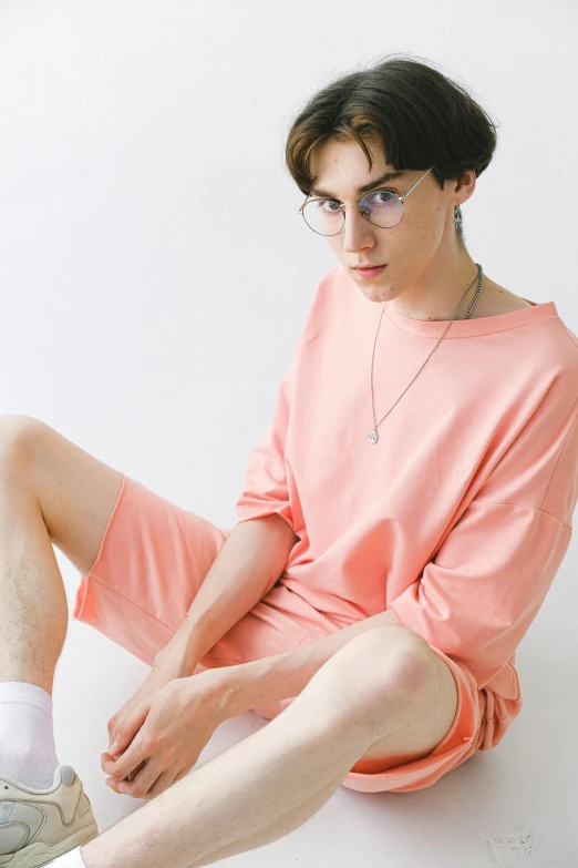 a man sitting on the ground with his legs crossed, a picture, by Eizan Kikukawa, unsplash, neo-dada, in shades of peach, wearing a baggy pajamas, androgynous male, dolman