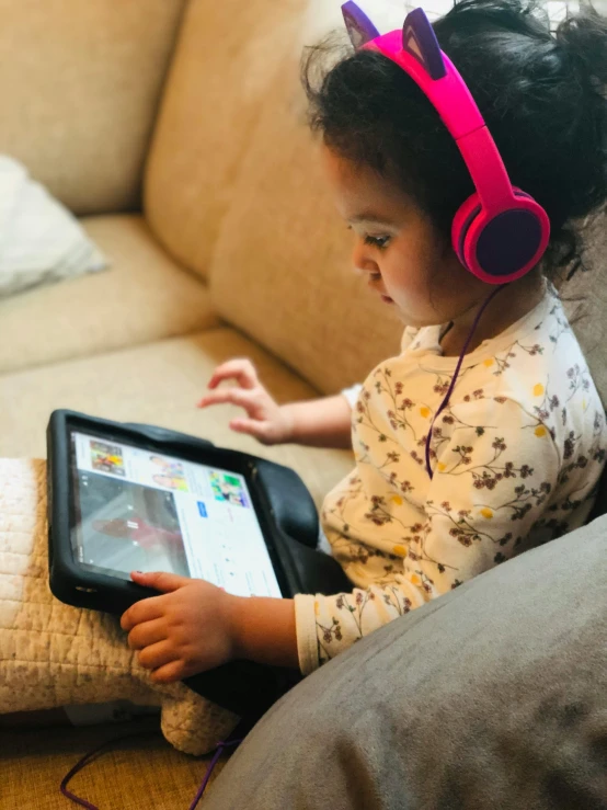 a little girl sitting on a couch using a tablet computer, an album cover, pexels contest winner, happening, pink headphones, profile picture 1024px, looking towards camera, wearing headset