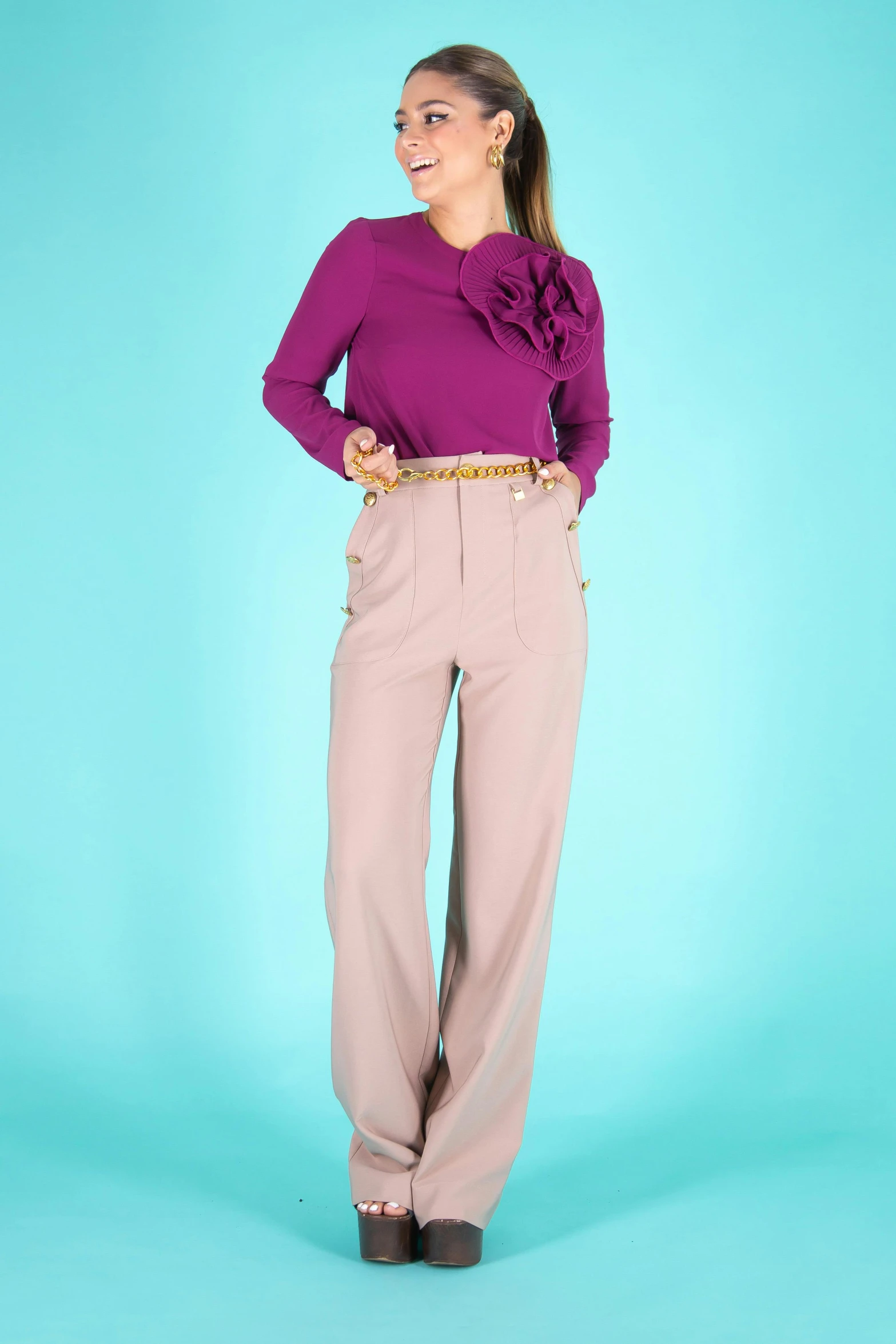 a woman standing in front of a blue background, a colorized photo, inspired by Violet Fuller, brown pants, lookbook, pink and gold, productphoto