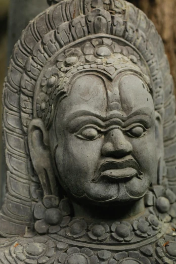 a close up of a statue with a tree in the background, south east asian with round face, ornate headdress, grey, scowling