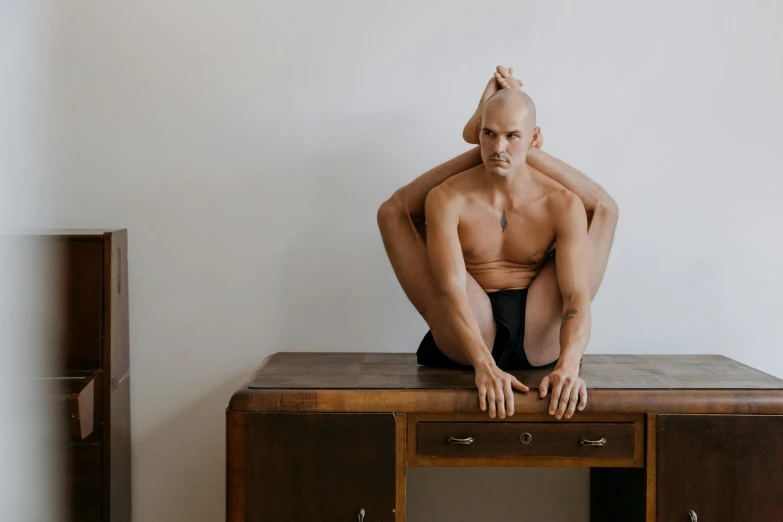 a man sitting on top of a wooden table, by Emma Andijewska, unsplash, renaissance, hairless, heroic shooting bow pose, yoga, hugging his knees