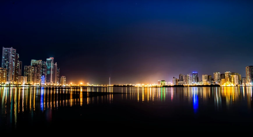 a large body of water next to a city at night, by Ibrahim Kodra, pexels contest winner, hurufiyya, long beach background, karachi skyline background, neon reflections, panoramic