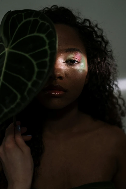 a woman holding a green leaf in front of her face, an album cover, by Lily Delissa Joseph, trending on pexels, afrofuturism, glowing in the dark, iridescent image lighting, sensual lighting, portrait of vanessa morgan