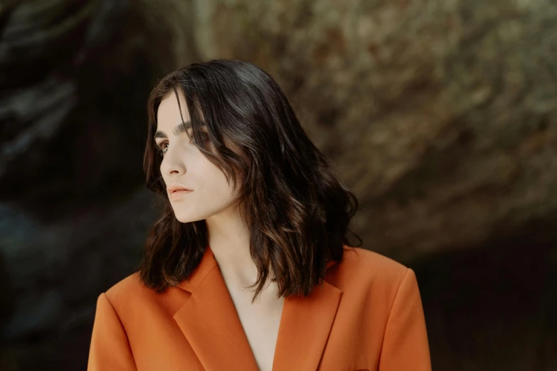 a woman in an orange blazer standing in front of a rock, an album cover, trending on pexels, photorealism, arya stark, rachel weisz, alessio albi, handsome girl