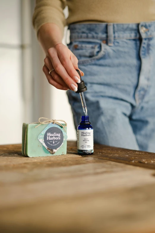 a person with a bottle of essential oil on a table, by Helen Stevenson, happening, textured base ; product photos, holding syringe, label, handcrafted