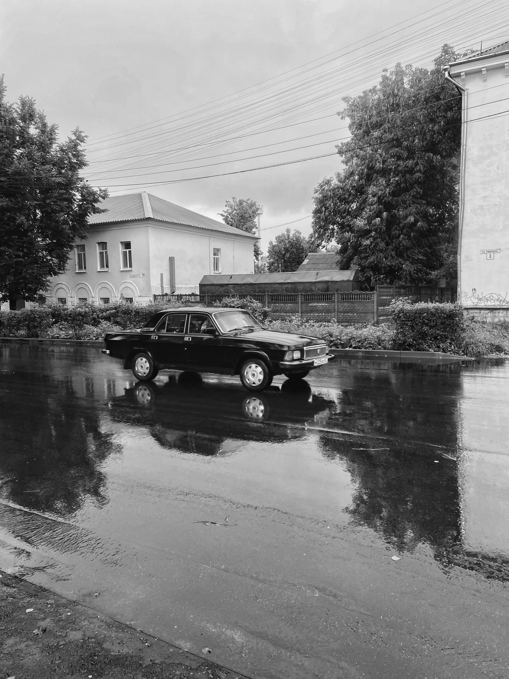 a black and white photo of a car in the rain, a black and white photo, by Sergei Sviatchenko, soviet town, water reflection!!!!!, analogue photo low quality, photo on iphone