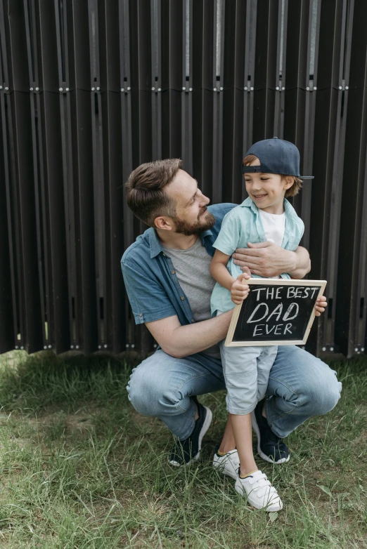 a man holding a sign that says the best dad ever, by Julia Pishtar, pexels contest winner, panel, denim, wholesome, van