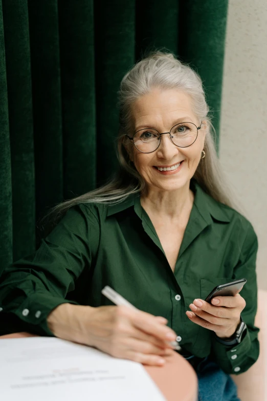 a woman sitting at a table with a cell phone, a character portrait, pexels contest winner, straight grey hair, lush green, holding notebook, smiling confidently