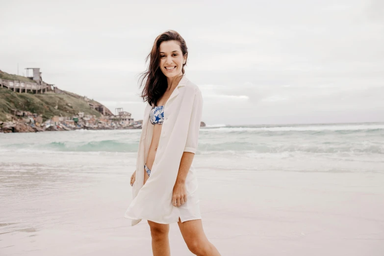 a woman standing on a beach next to the ocean, dirty linen robes, in front of white back drop, caio santos, looking happy
