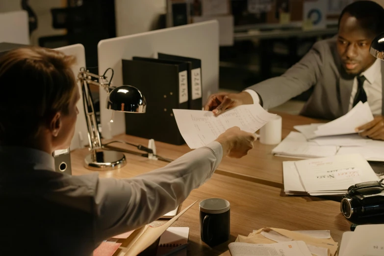 a couple of people sitting at a table with papers, on a desk, reaching out to each other, low lighting, in an office