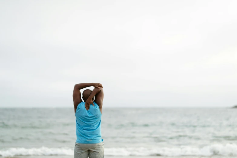 a man standing on top of a sandy beach next to the ocean, a picture, unsplash, figuration libre, wearing fitness gear, plain stretching into distance, relaxed. blue background, back turned