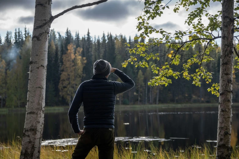 a man is taking a picture of a lake, inspired by Eero Järnefelt, foliage clothing, looking away from camera, in an open forest, explorer