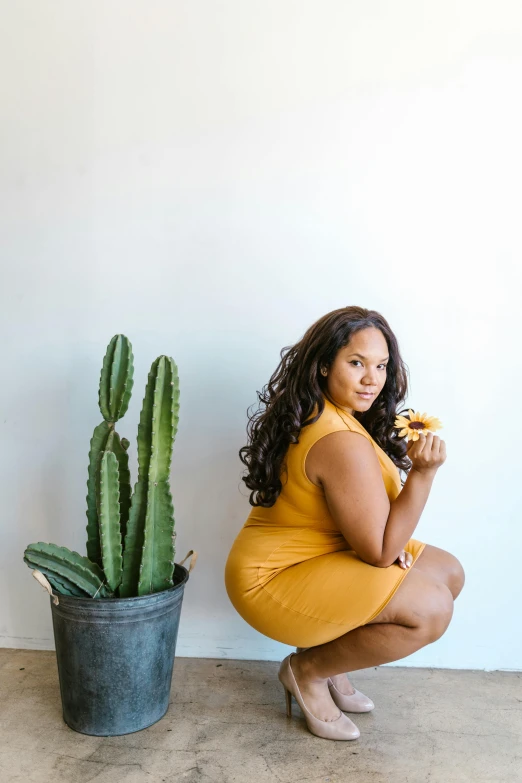a woman in a yellow dress squatting next to a cactus, a portrait, by Kristin Nelson, unsplash, eating a donut, full figured, with brown skin, studio picture