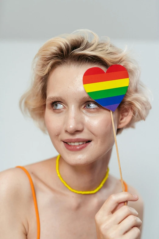 a woman holding a rainbow heart on a stick, trending on pexels, bauhaus, androgynous face, digital image, pale skin curly blond hair, photo booth