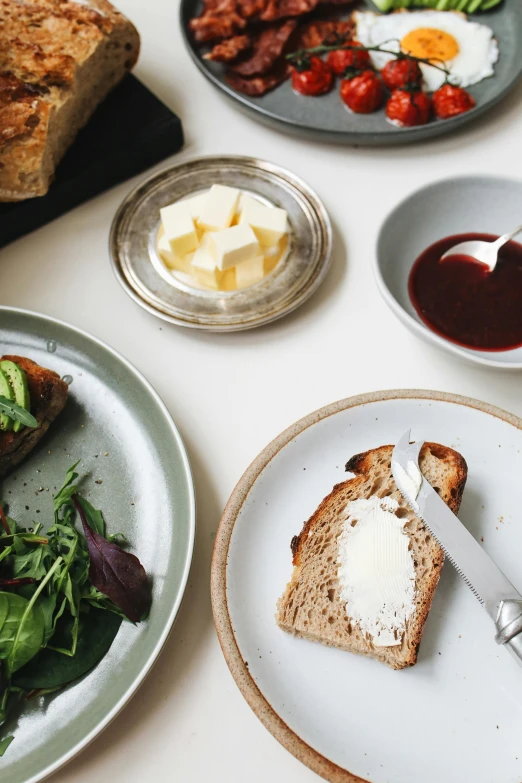 a white table topped with plates of food, a still life, inspired by Richmond Barthé, unsplash, brown bread with sliced salo, butter, grey, manuka