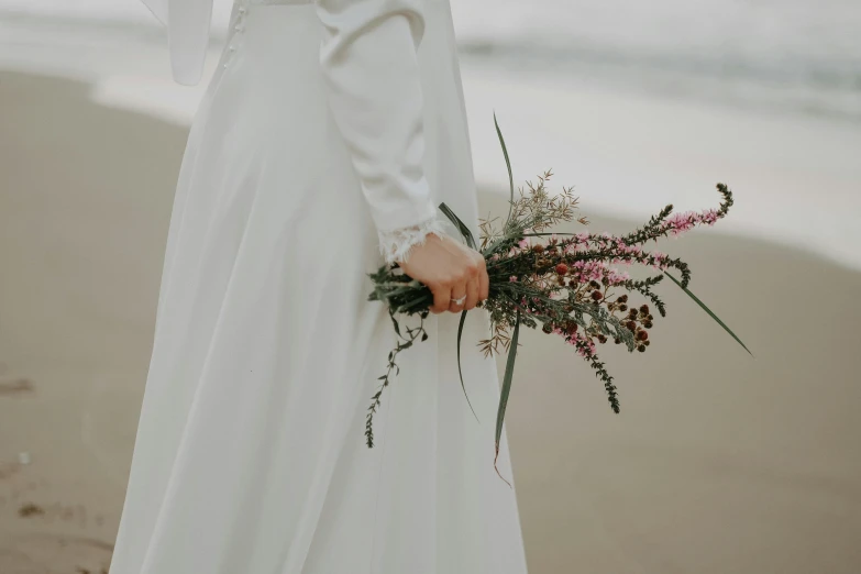 a woman standing on a beach holding a bouquet, unsplash, in a flowing white tailcoat, background image, detail shot, herbs and flowers