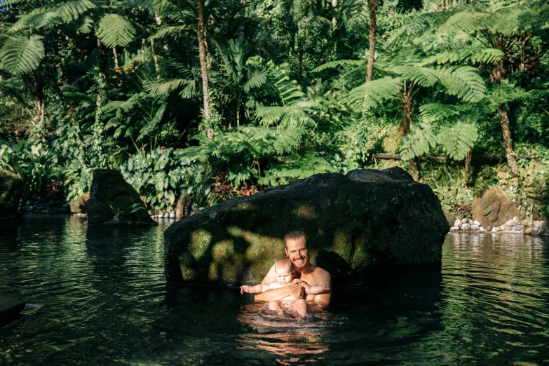 a man holding a baby in a body of water, sumatraism, above lush garden and hot spring, alana fletcher, rock pools, jen atkin