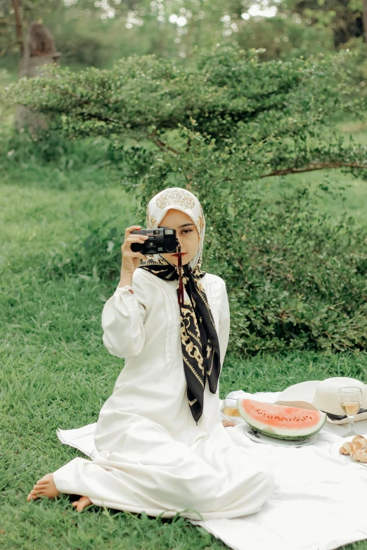 a woman sitting in the grass taking a picture with a camera, by Basuki Abdullah, white hijab, having a picnic, in garden, 8 k )