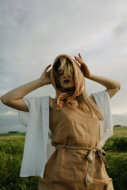 a woman standing in a field with her hands on her head, inspired by Andrei Rublev, trending on pexels, draped in silk, wearing farm clothes, with a covered face, young blonde woman