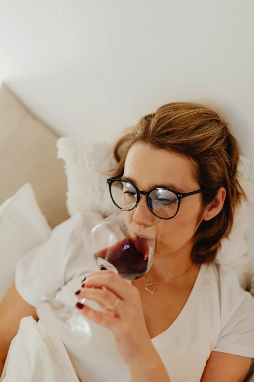 a woman sitting on a bed drinking a glass of wine, wearing glack glasses, up close, bedhead, glasses |