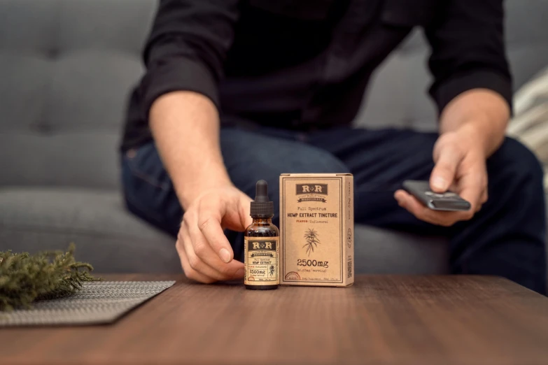 a man sitting on a couch holding a bottle of essential oil, a portrait, by Daniel Lieske, trending on pexels, renaissance, cigarrette boxes at the table, 3 d product render, bee, professional product shot