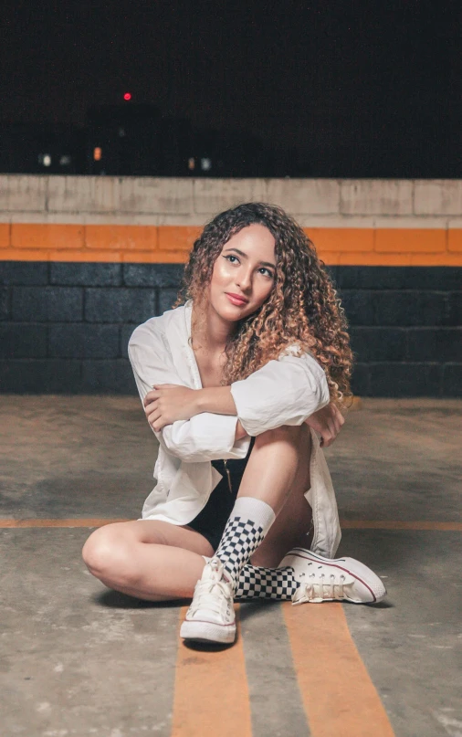 a woman sitting on the ground in a parking garage, a portrait, inspired by Amelia Peláez, trending on pexels, teenage vanessa morgan, curly haired, wearing white sneakers, concert photo
