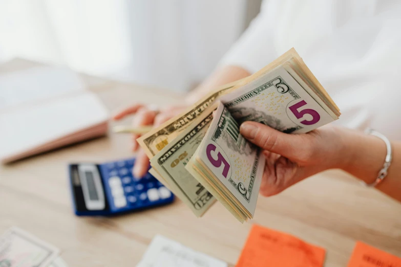 a person holding a stack of money in front of a calculator, pexels contest winner, 🦩🪐🐞👩🏻🦳, back of hand on the table, avatar image, full product shot