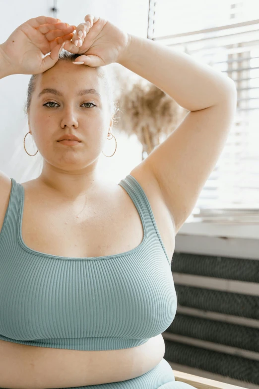 a woman sitting in front of a window with her hands on her head, a colorized photo, inspired by Elsa Bleda, trending on pexels, renaissance, she has a jiggly fat round belly, wearing : tanktop, wearing a light blue shirt, looking confident