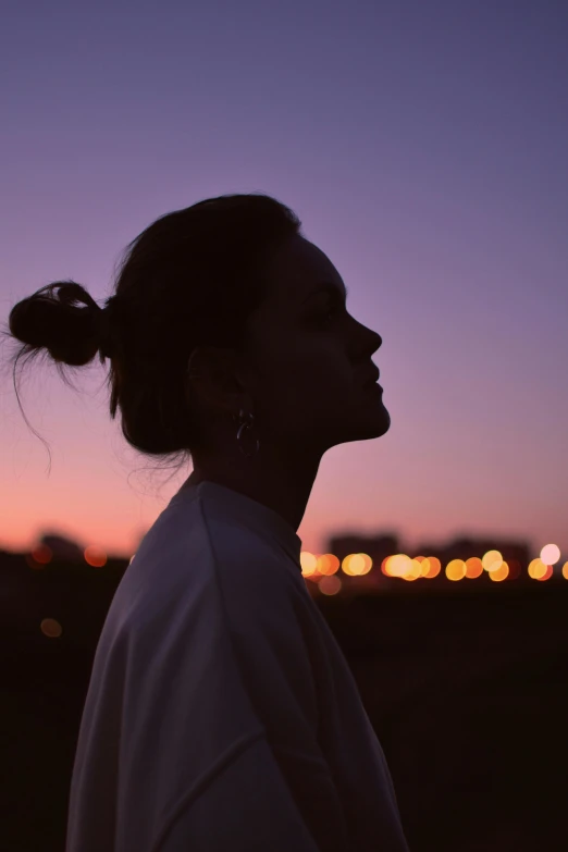 a woman standing on top of a grass covered field, a picture, trending on pexels, city lights in the background, prominent jawline, pastel palette silhouette, girl with messy bun hairstyle