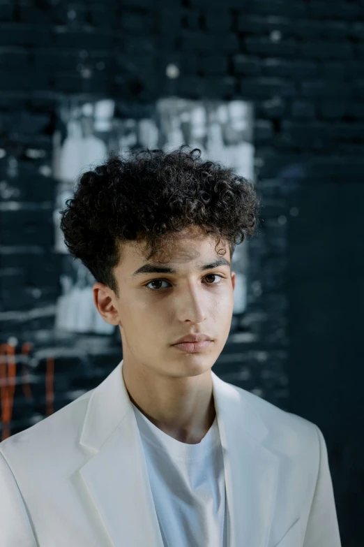 a man in a white suit standing in front of a brick wall, an album cover, inspired by Alexis Grimou, trending on pexels, renaissance, (dark shorter curly hair), 14 yo berber boy, portrait. 8 k high definition, androgynous face