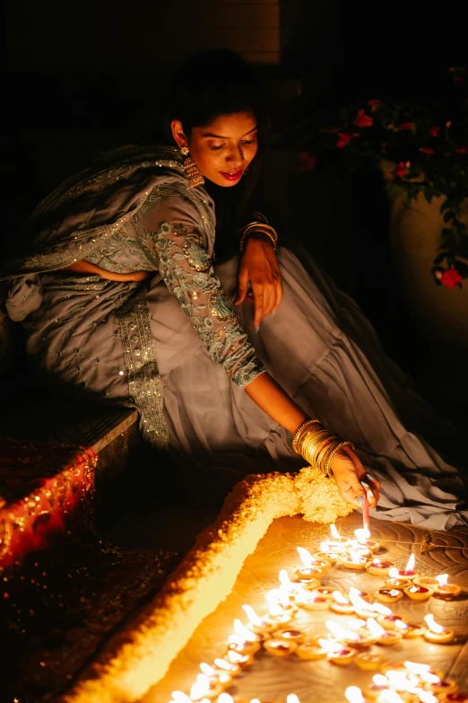 a woman sitting on the ground lighting candles on a cake, pexels contest winner, visual art, wearing traditional garb, grey, lights with bloom, fire pit